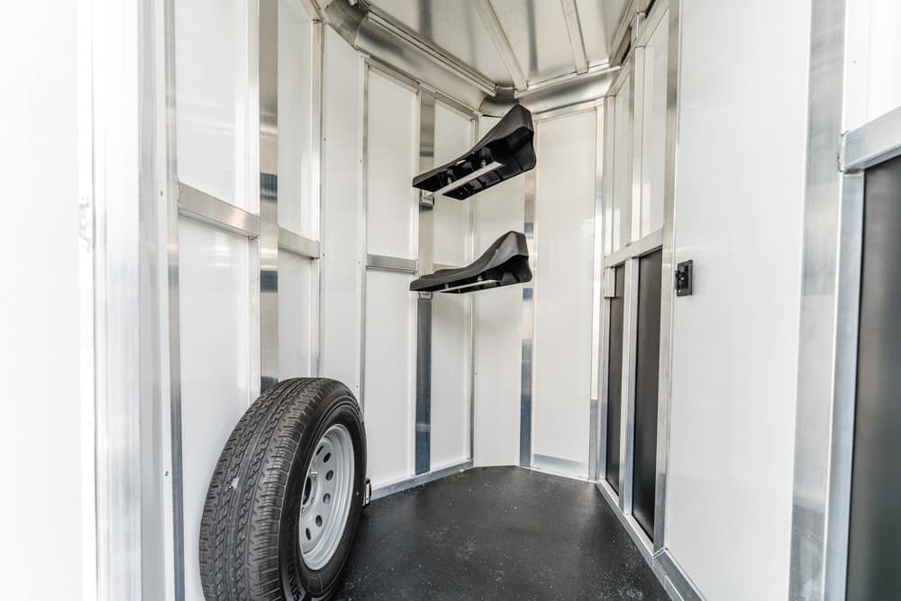 Inside of a white trailer with a spare tire leaning against the wall and two black wall-mounted racks/saddles above it.