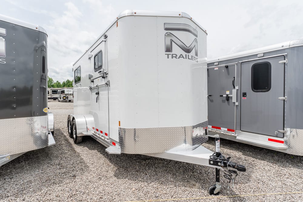 White enclosed gooseneck trailer with diamond-plate front, parked between gray trailers on a gravel lot at a dealership/parking lot.