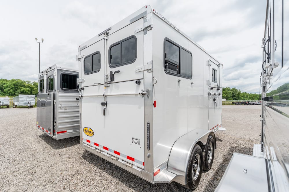 White aluminum enclosed trailer with two axles and rear double doors, parked on a gravel lot with other trailers nearby.