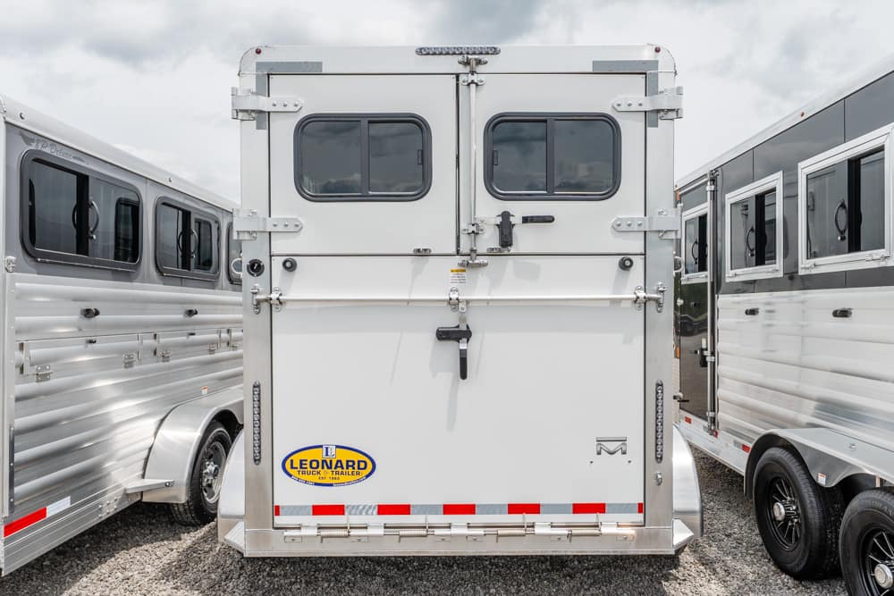 Rear view of a white horse trailer with two small rear doors, safety latches, and a Leonard trailer logo, parked between aluminum trailers in a lot.