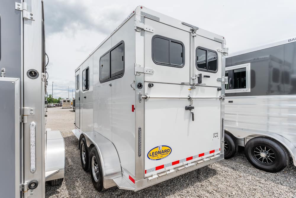 White two-axle horse trailer with rear double doors, small windows, and a Leonard logo, on a gravel lot.