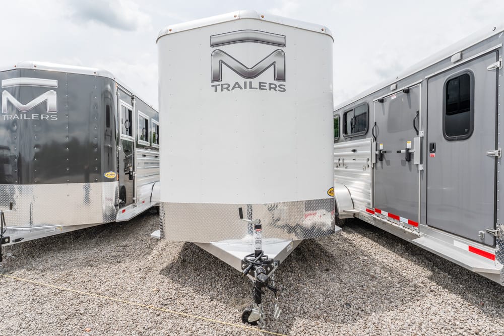 White front trailer with M Trailers logo between two gray trailers on a gravel lot; tow hitch and jack visible at front.