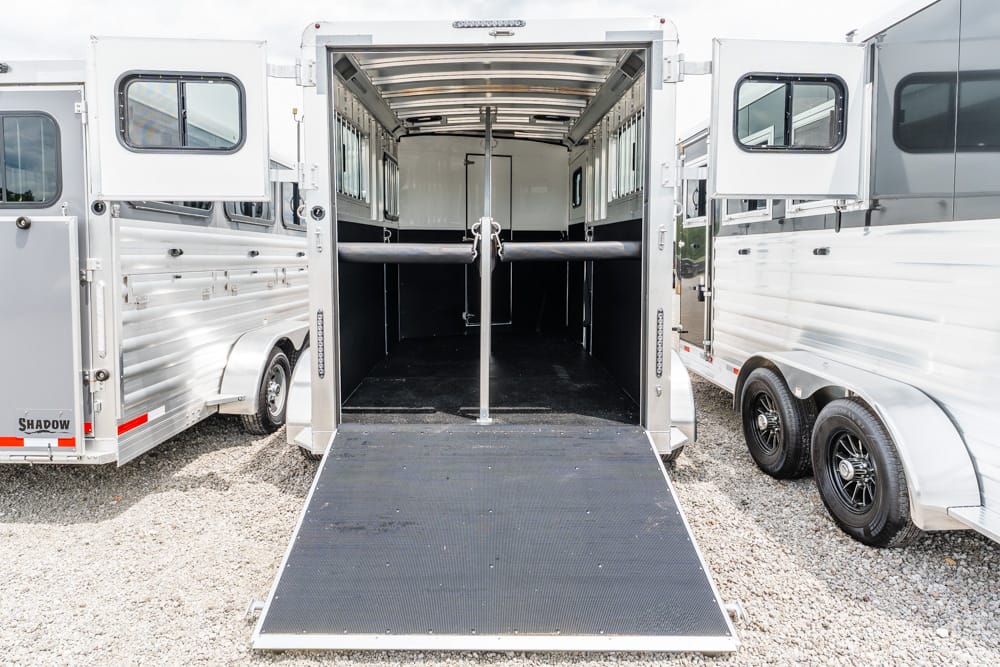 Rear view of a horse trailer with the ramp down, showing stalls and a center partition inside a row of aluminum trailers on a gravel lot