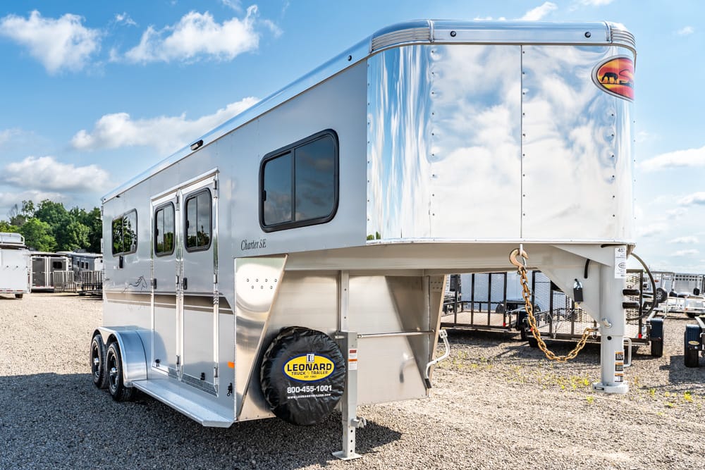 Silver gooseneck travel trailer with living quarters parked on a gravel lot; front has a chrome front cap and a spare tire cover reading Leonard Truck & Trailer.