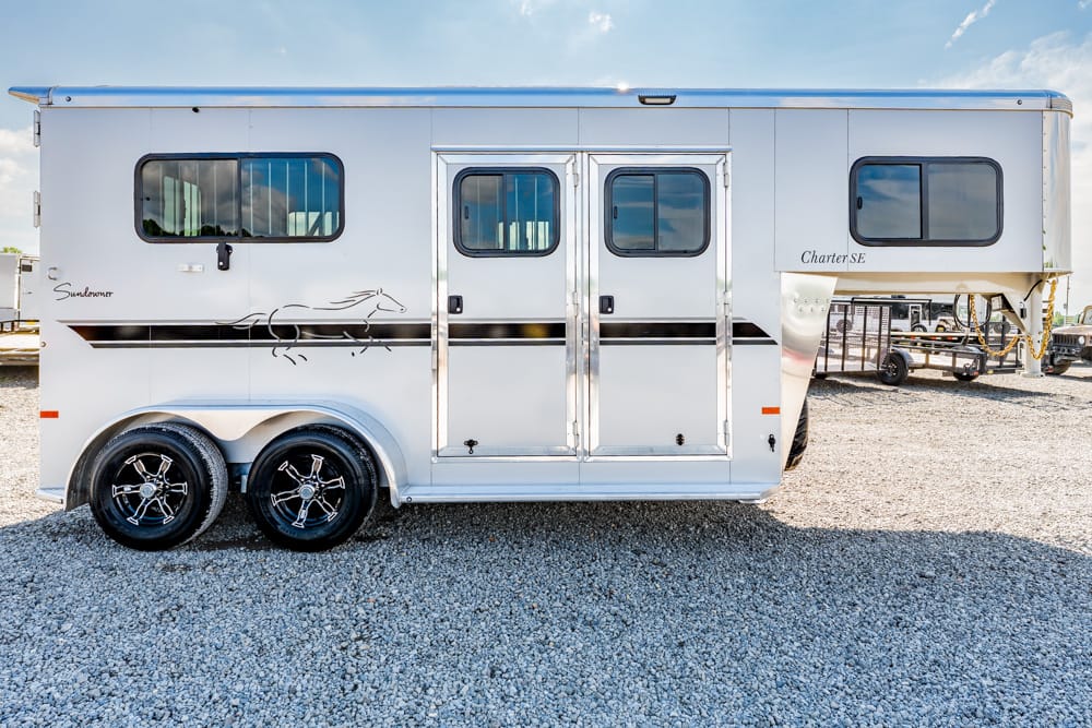 White Sundowner horse trailer with dual axles, side windows, and a decorative horse outline on a gravel lot