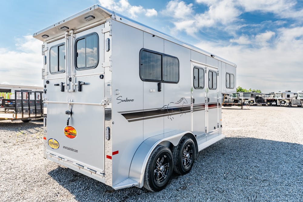 Silver Sundowner horse trailer with two axles parked on a gravel lot under a blue sky, showing side and rear doors and windows.