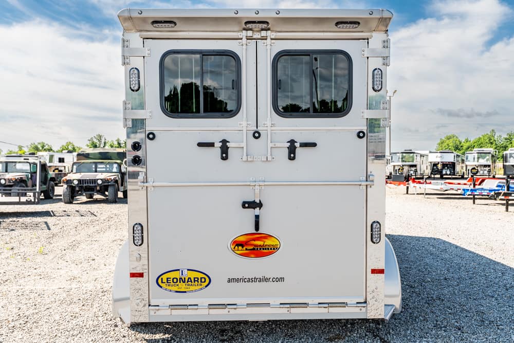 Rear view of a white trailer with two rounded windows, metal latches, and branding logos on the back (Leonard Trailer, Sundowner) in a gravel lot.