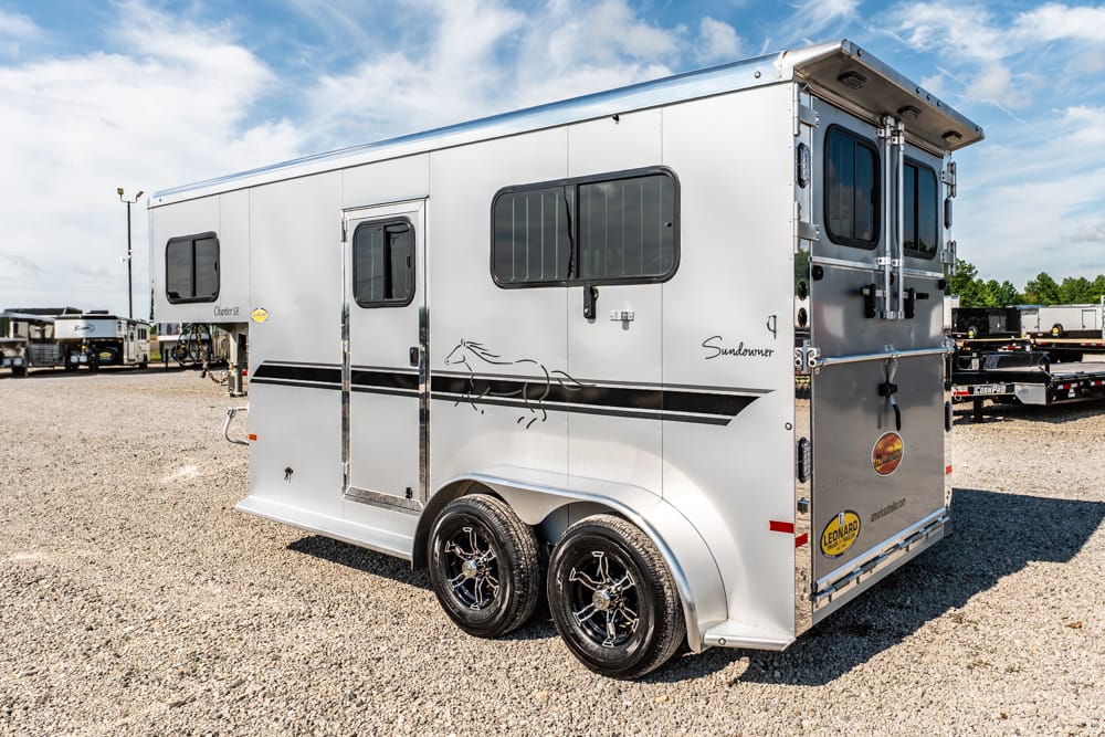Silver Sundowner horse trailer with dual axles and black stripe parked on a gravel lot, windows visible on the side
