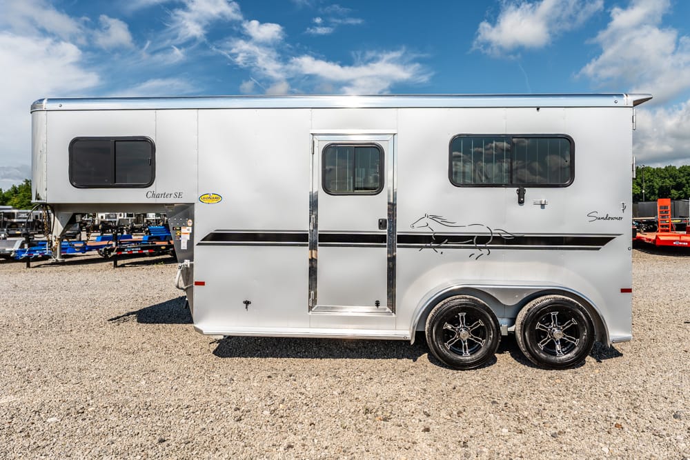 White aluminum horse trailer with two axles, chrome trim, and a decorative horse graphic along the side, parked on a gravel lot under a blue sky.