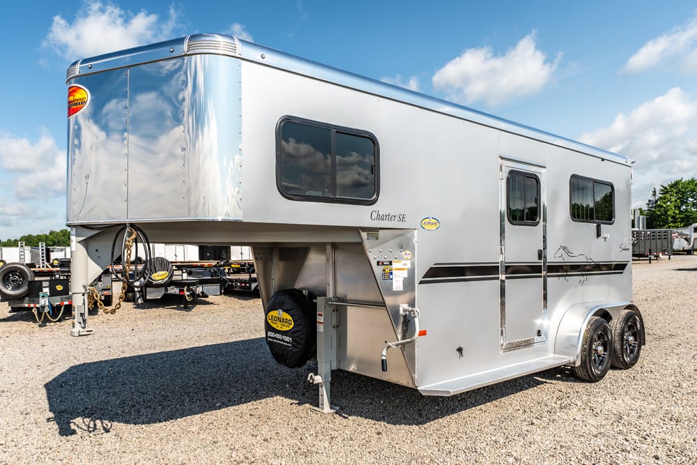 Silver gooseneck fifth-wheel travel trailer parked on a gravel lot, with two axles, spare tire, and side entry door under a clear blue sky.