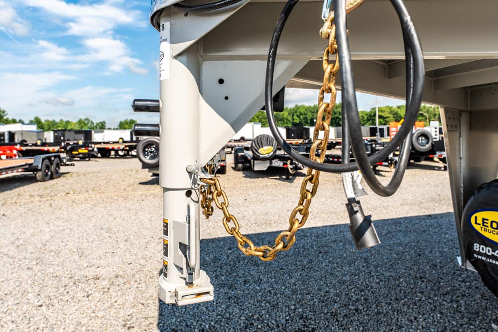 Close-up of a trailer hitch with a gold safety chain and black electrical cable on a gravel lot with other trailers in the background.