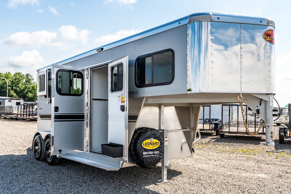 Silver two-axle gooseneck horse trailer with open side door and a spare tire cover, parked on gravel under a blue sky.