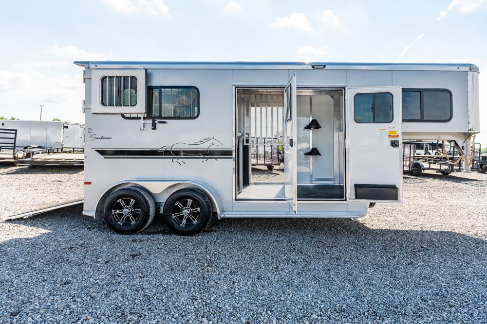 White horse trailer with two axles parked on gravel; side and rear doors open, showing interior stalls and tack area.