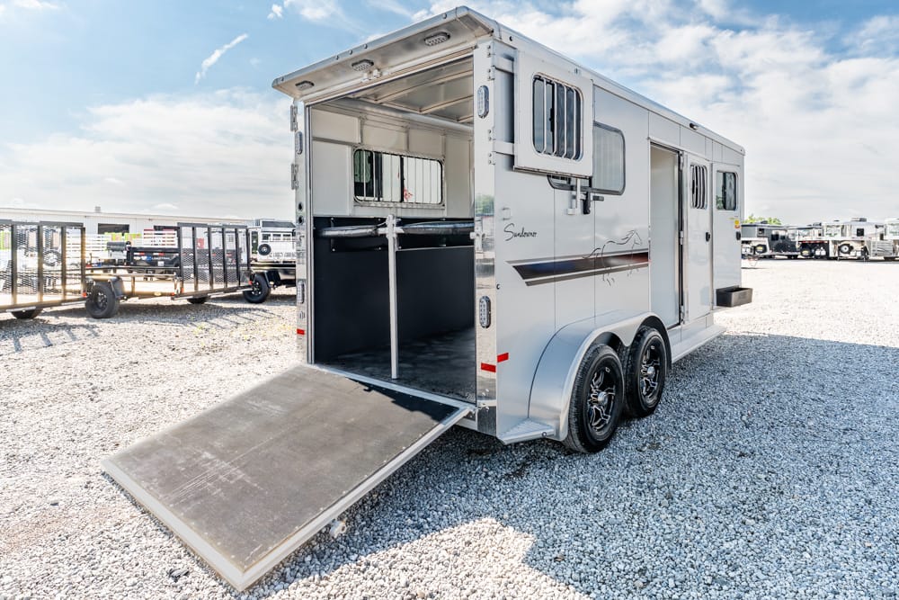 Silver horse trailer with open rear ramp and divider, parked on gravel with other trailers in the background.