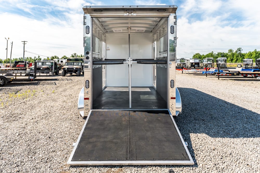 Open aluminum trailer with ramp down, empty interior with a central divider, parked on a gravel lot while other trailers line up in the background.