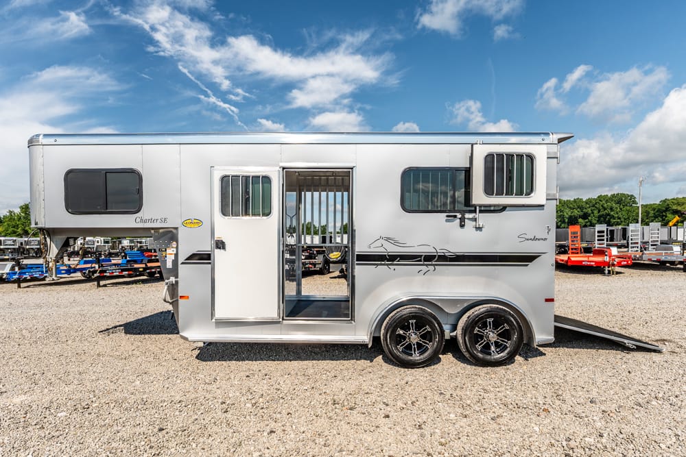 Silver horse trailer with two axles parked on gravel; side door open and rear ramp lowered, showing interior access for horses or gear.