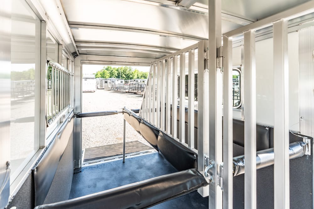 Interior of a metal animal transport trailer with vertical dividers and black padded benches, sunlight at the rear opening.
