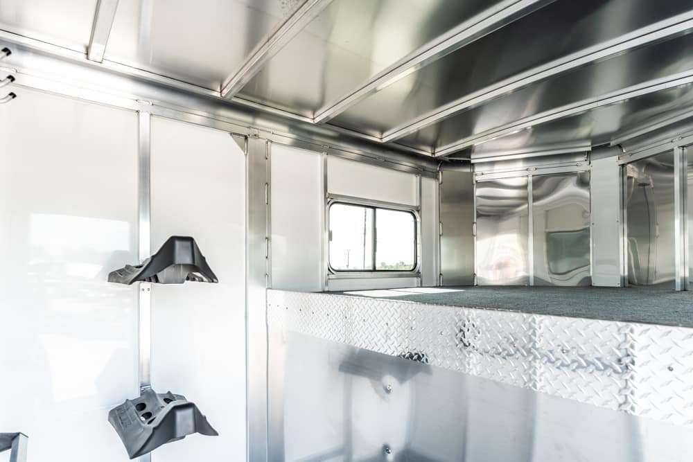 Interior of a shiny aluminum trailer with diamond-plate flooring and a small side window, currently empty and well-lit.