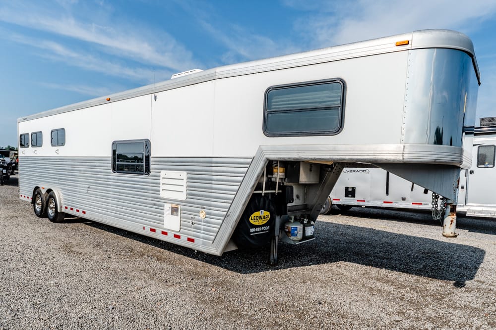 Silver gooseneck horse trailer on a gravel lot, with windows, dual axles, and a spare tire mounted at the front.