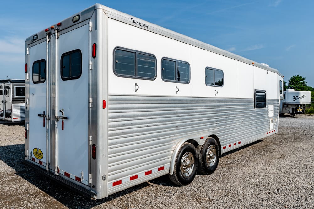 Silver horse trailer with side windows and tandem wheels on a gravel lot under a blue sky