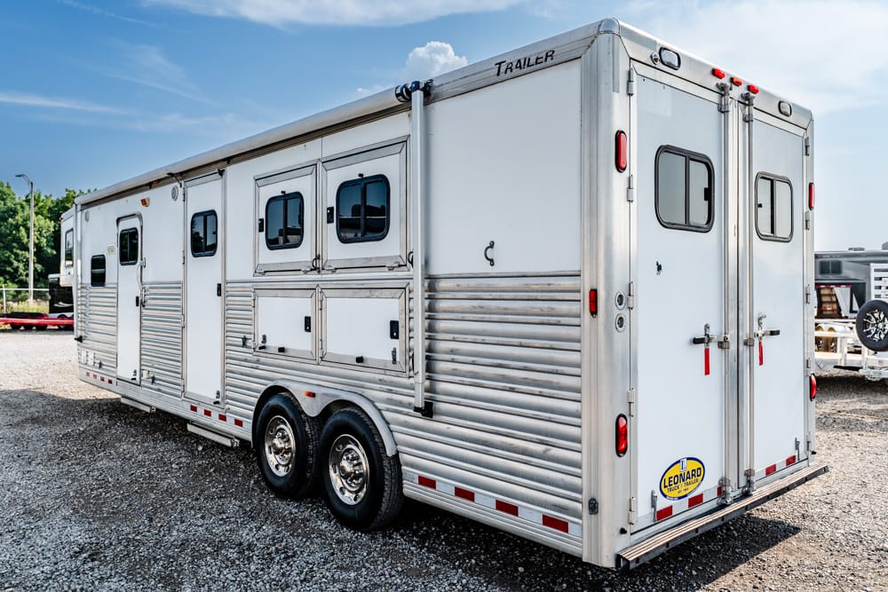 Silver aluminum horse trailer with dual axles parked on gravel, side compartments visible, rear doors closed, clear blue sky above