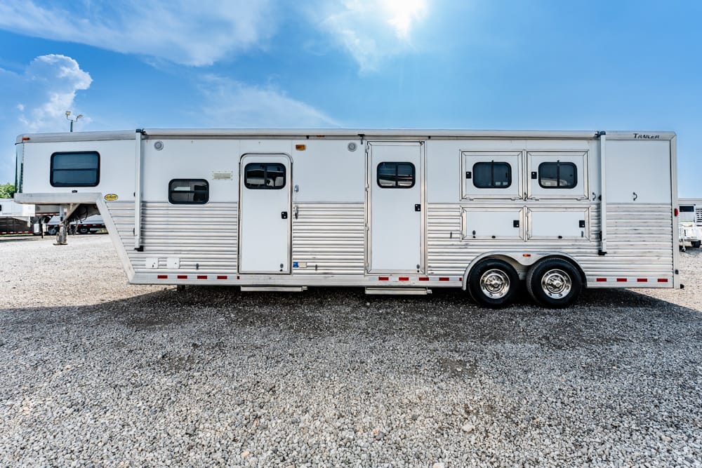 Long silver horse trailer with multiple stall doors parked on a gravel lot under a blue sky.