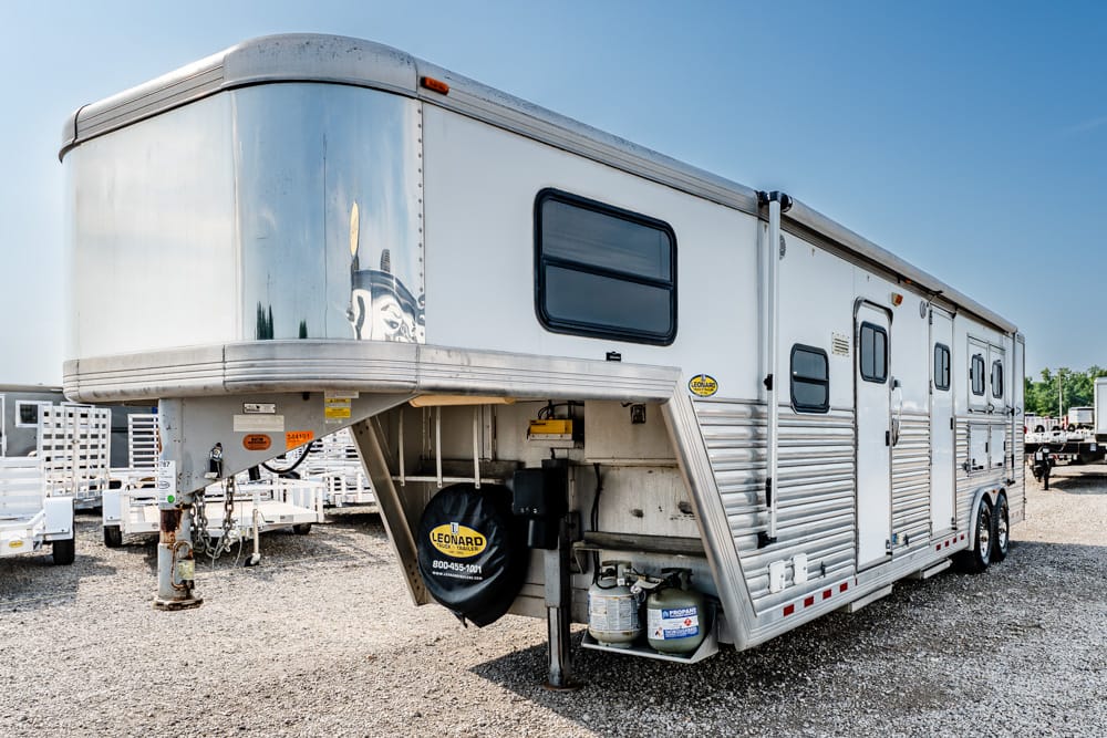 Silver fifth-wheel trailer parked on a gravel lot, showing its front hitch, spare tire cover, propane tanks, and multiple windows and doors on the side.