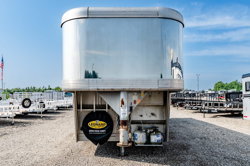 Silver rounded-top trailer with spare tire cover and propane cylinders at the hitch, parked in a gravel trailer yard.