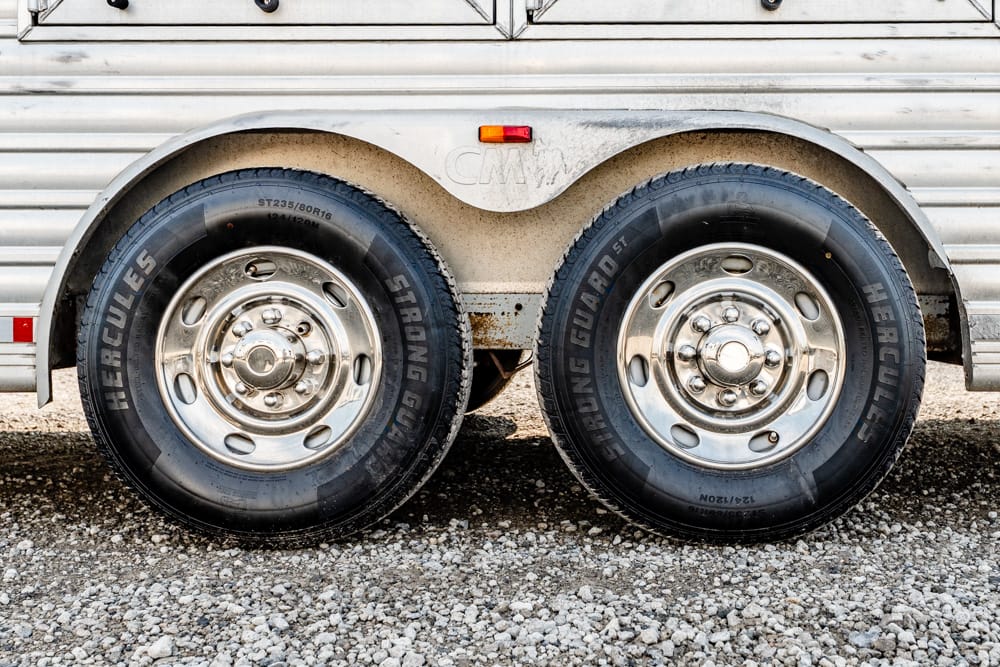 Twin trailer wheels with chrome hubs on a silver aluminum trailer, resting on gravel.