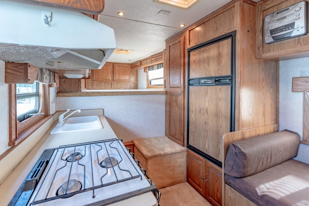 Compact RV kitchen with a three-burner stove, sink, and wooden cabinets; fridge to the right and a small seating area nearby