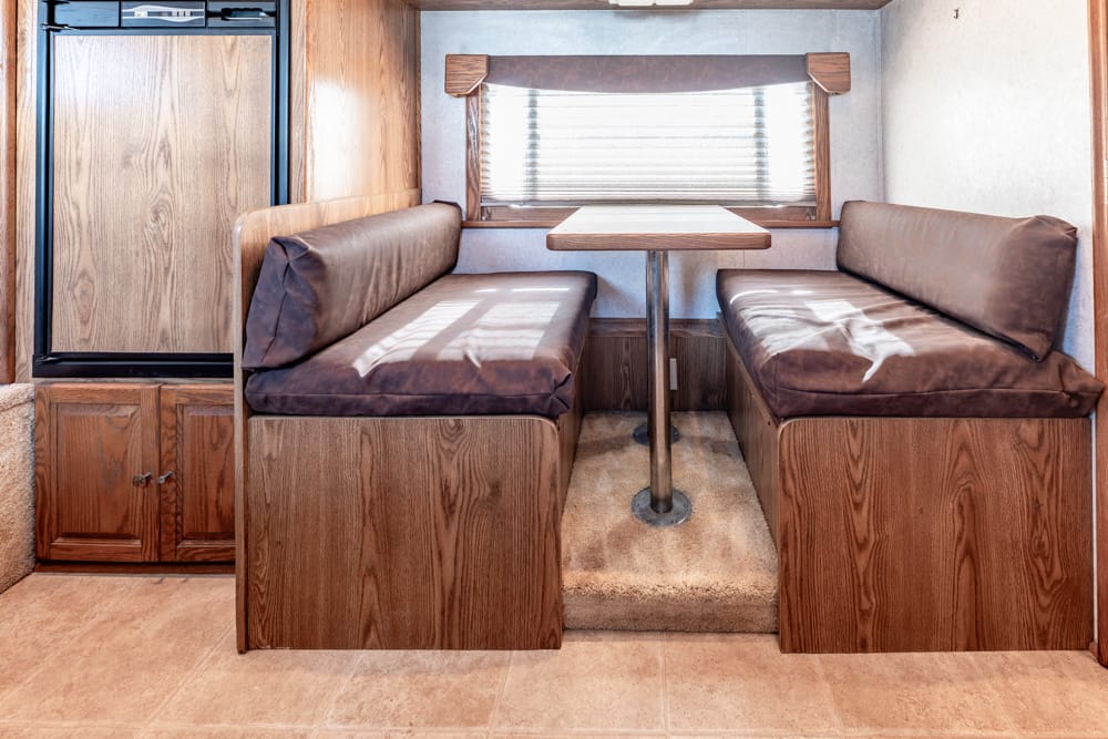 RV dinette with two brown upholstered benches and a wooden table in between, under a window with blinds.