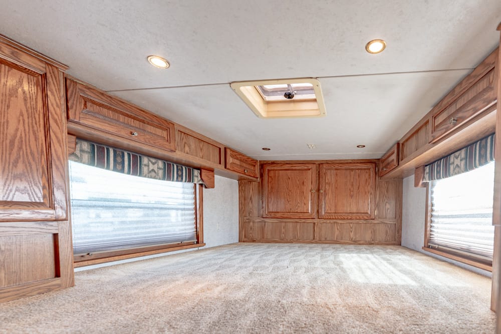 RV bedroom with oak cabinets along both sides, carpeted floor, and daylight from side windows with blinds.