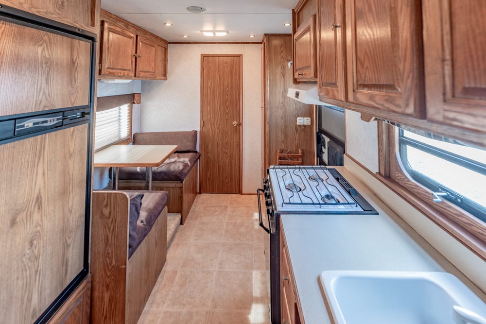 Interior of a compact RV kitchen: wooden cabinets, a stove on the right, and a dinette with bench seating on the left under a window.
