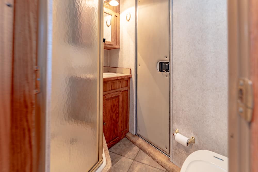 Small RV bathroom with a wooden vanity, sink, and frosted shower door along a narrow aisle.