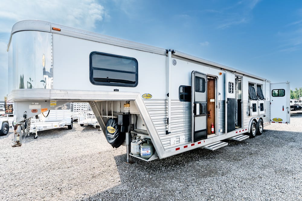 Large white travel trailer with multiple entry doors and metal steps, parked on a gravel lot under a blue sky.