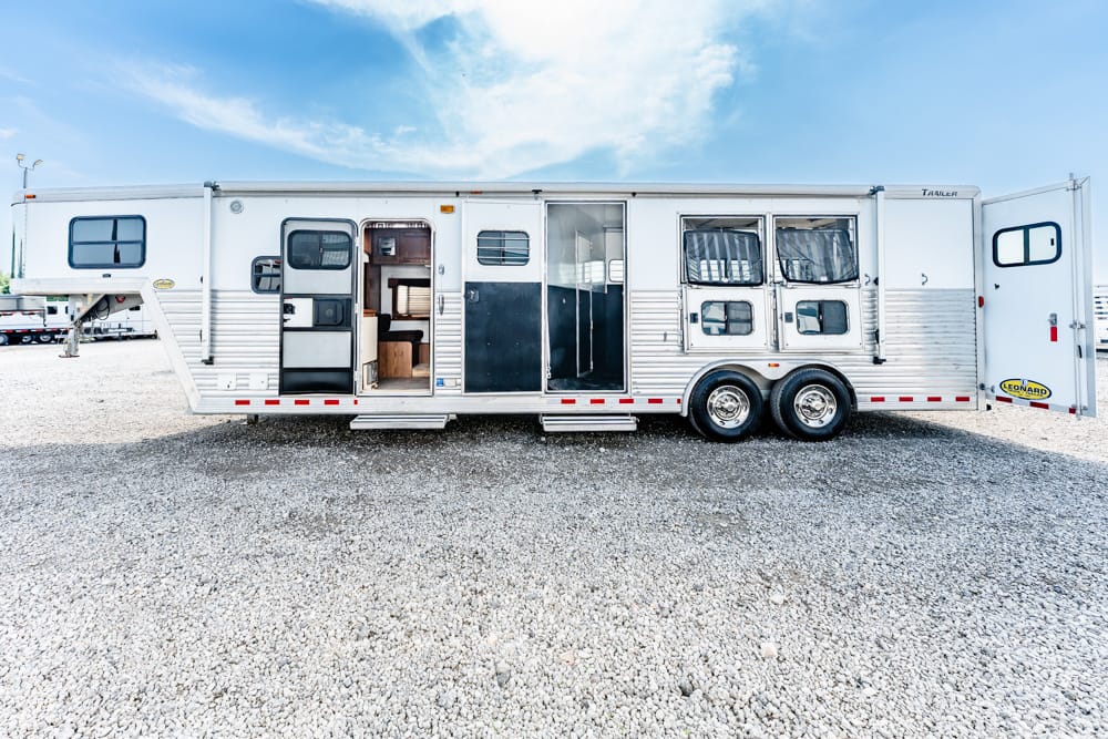 Long white aluminum trailer with living quarters, doors open to show interior steps and seating, parked on a gravel lot under a blue sky