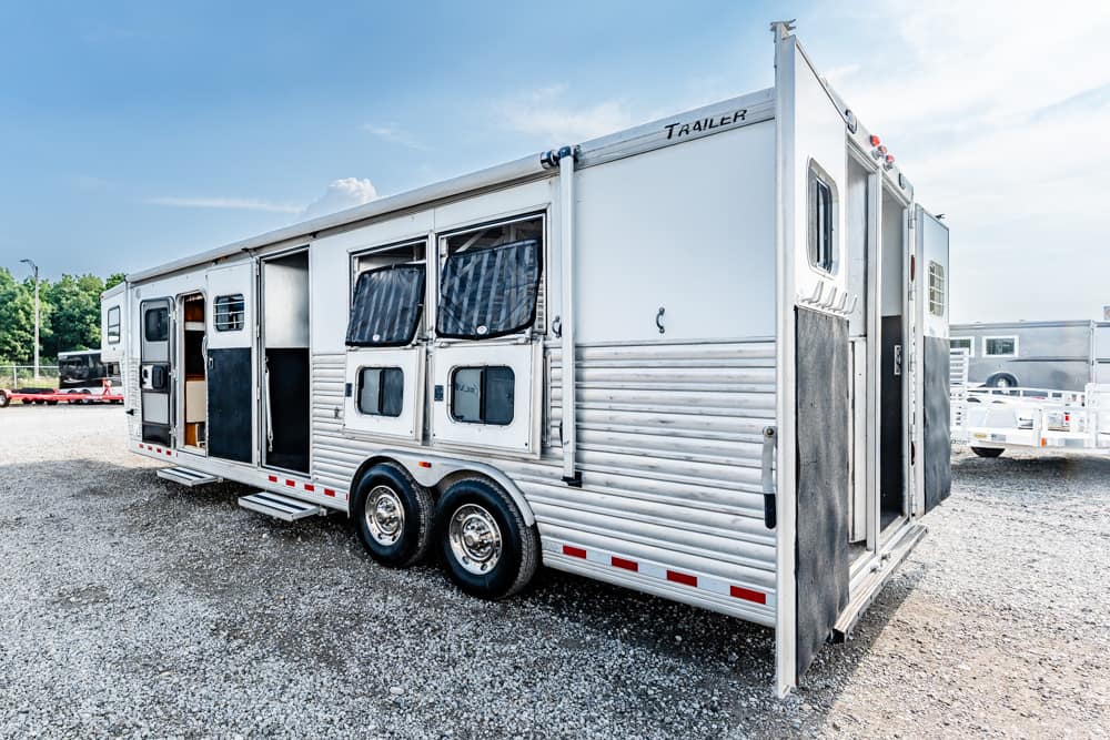A long silver horse trailer with multiple windows and open doors parked on a gravel lot, side-by-side wheels visible.