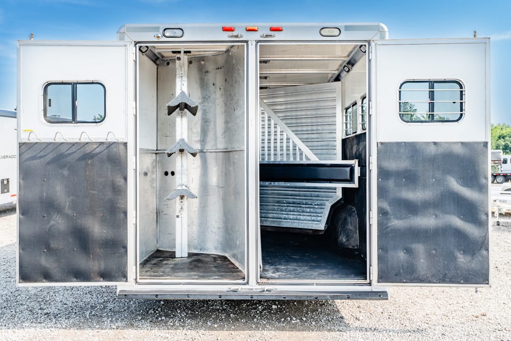 Open horse trailer with empty stalls and saddle racks inside, rear doors open on a gravel lot with a blue sky above