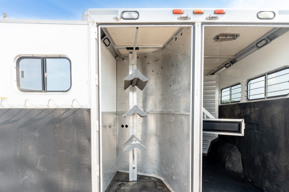 Interior of a horse trailer with a vertical metal tack rack featuring three angled holders mounted on a central post, and a sliding drawer visible to the right.