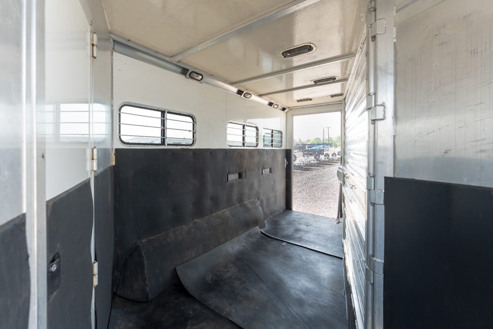 Interior of a horse trailer with a black padded divider and rubber mats, rear door open to a gravel lot outside.