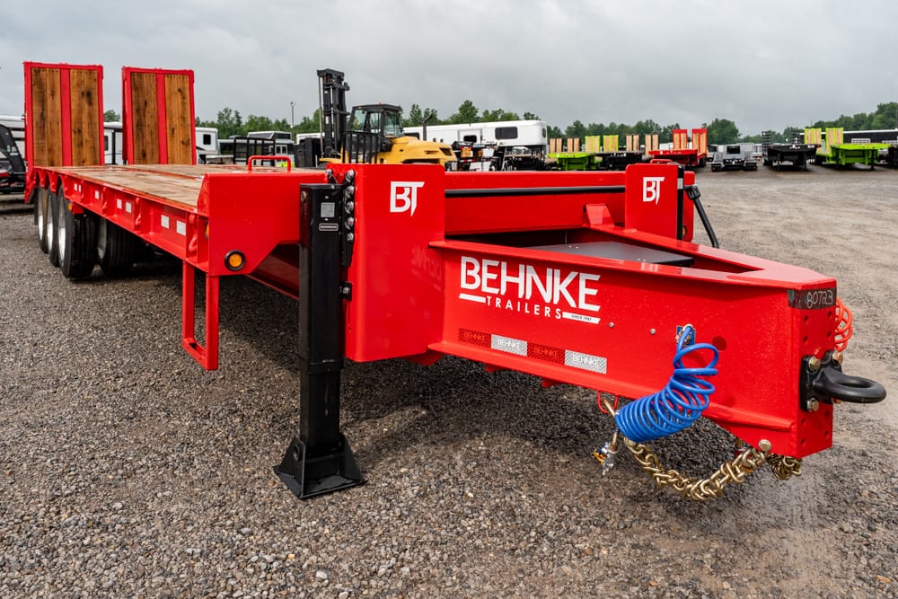 Red Behnke flatbed trailer with a blue coiled hose and safety chains in a gravel lot of trailers.