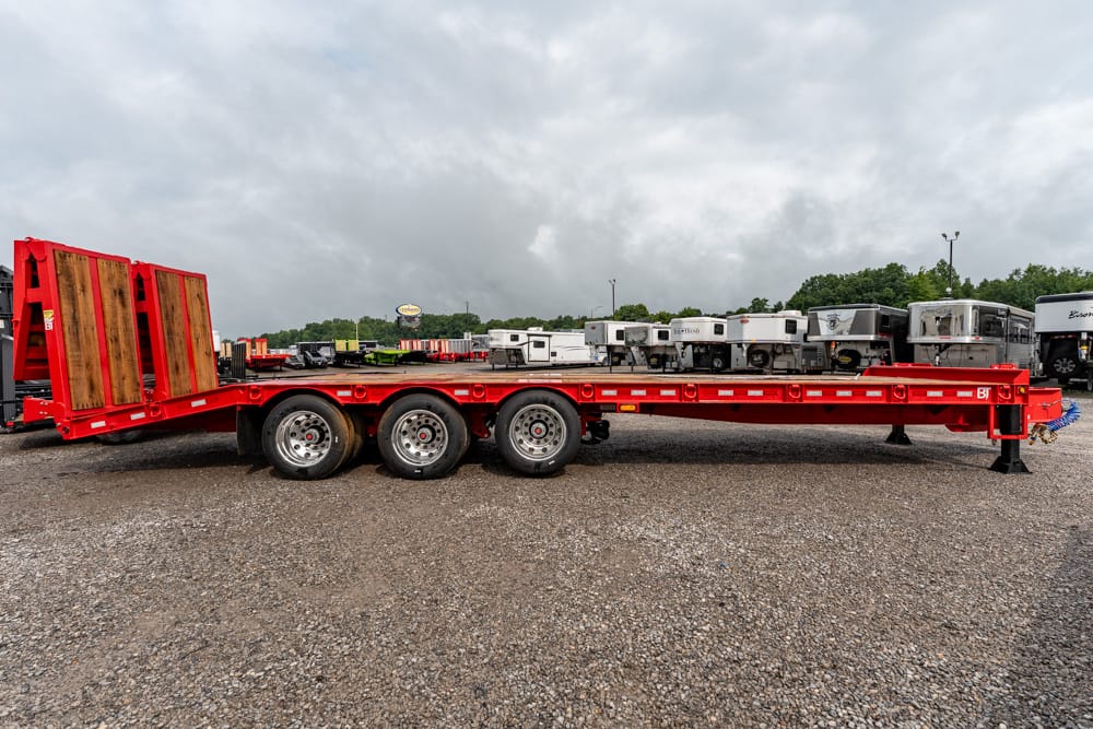 Red lowboy trailer with three axles and a wooden rear ramp, parked on a gravel lot in a truck yard.