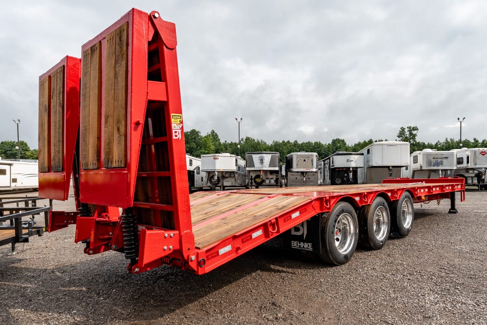 Red heavy‑duty flatbed trailer with fold-up wooden ramps raised at the back, triple axles, parked in a gravel lot with other trailers in the background