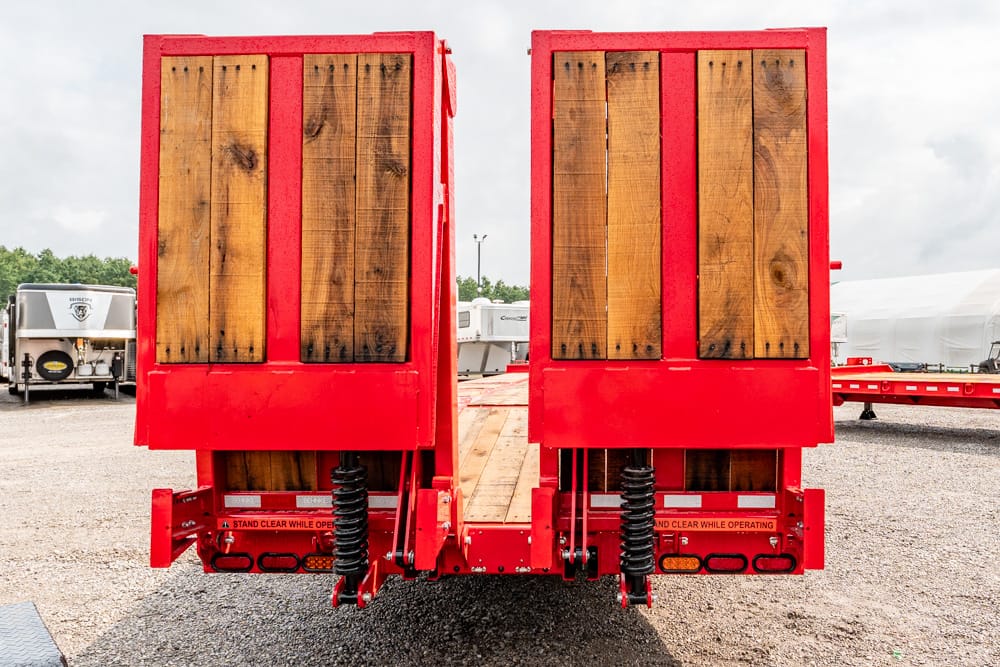 Back view of a red double-door flatbed trailer with wooden panels, parked on gravel lot, industrial setting in background.