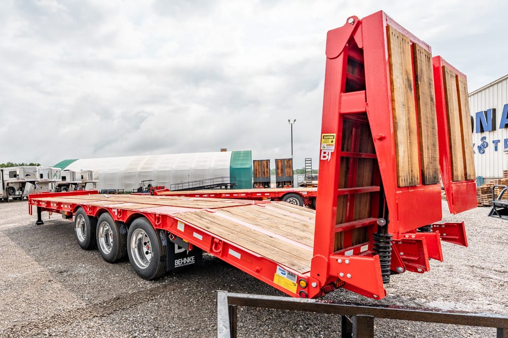 Red heavy-duty flatbed trailer with a wooden deck and triple-axle configuration in a gravel lot, ready for loading equipment.