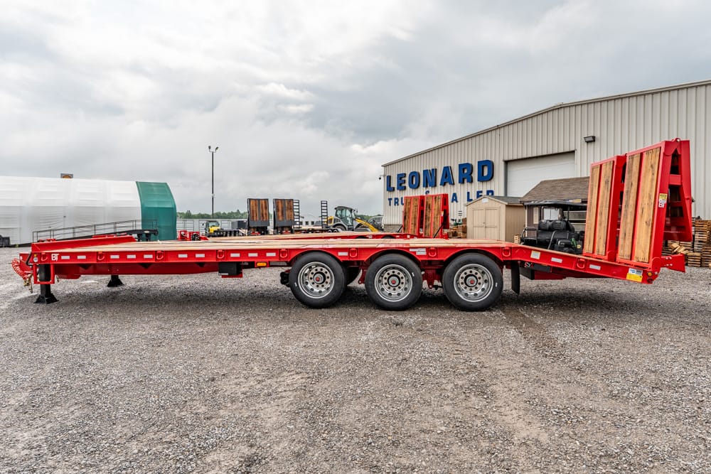 Red multi-axle flatbed trailer with loading ramps at the rear, parked on a gravel lot near an industrial building.