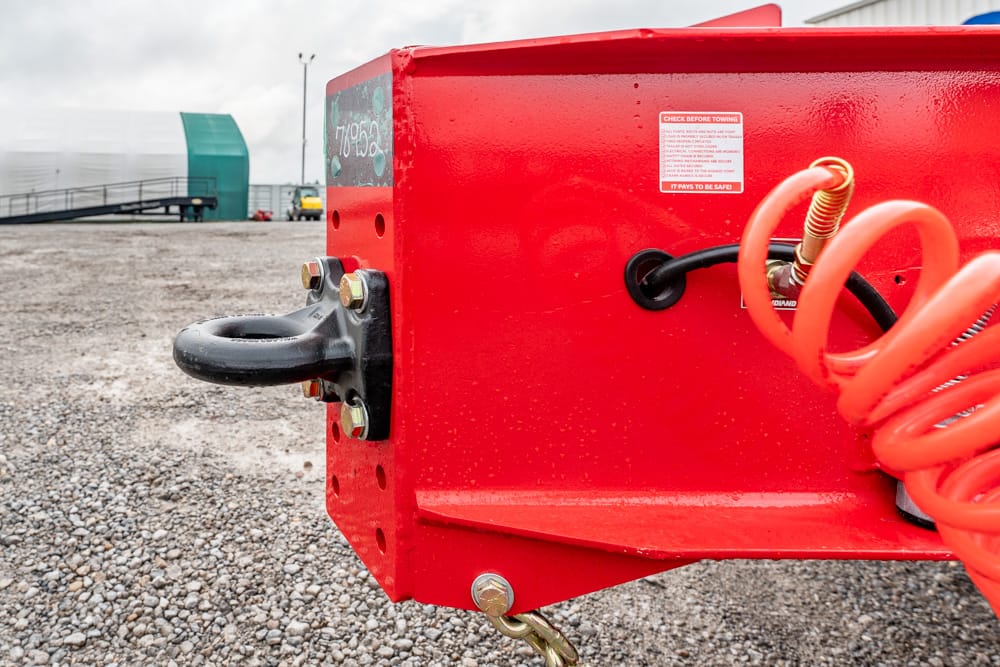 Red trailer frame with a black hitch ball and bright orange coiled air hose on a gravel lot.
