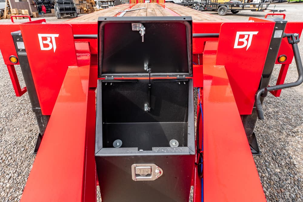 Red BT trailer with central open storage box revealing dark interior; gravel yard and equipment in background, indicating an industrial/maintenance setting.