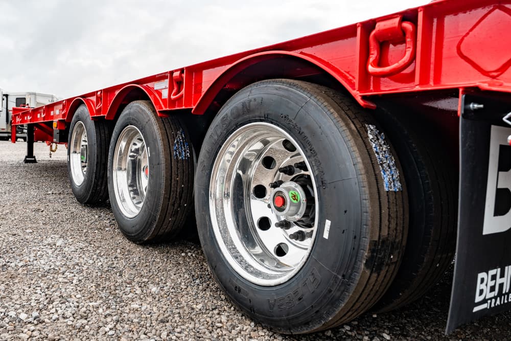Red flatbed trailer with triple axle and chrome wheels on a gravel lot, close view of tire treads and hub caps.