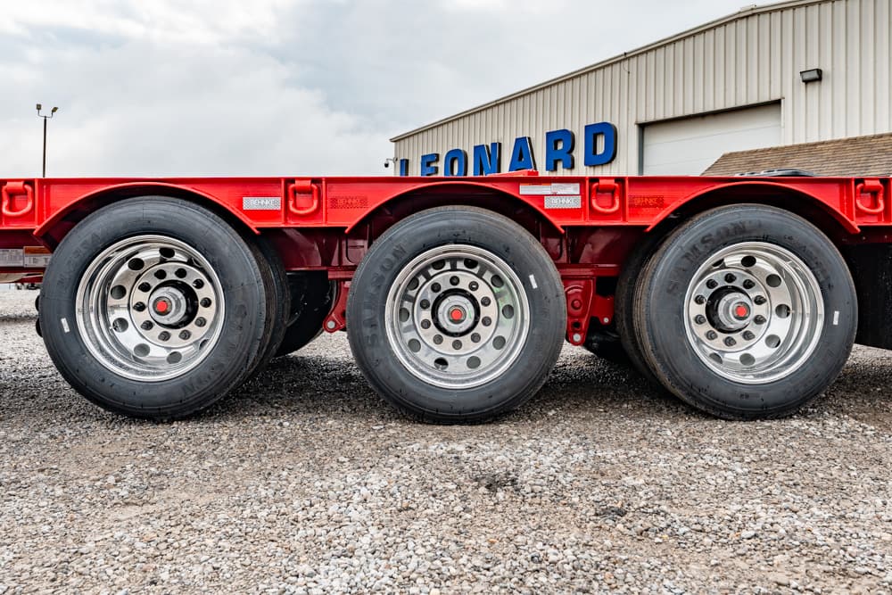 Close-up of a red flatbed trailer with three axles and chrome wheels on a gravel lot, an industrial building in the background with blue letters.